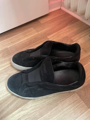 Svarta slip-on sneakers från Arigato - Stilrena svarta slip-on sneakers från Arigato med ovandel i mocka och elastiskt band över vristen. Tjock vit sula och diskret logga på sidan och tungan. Perfekta för en clean och modern look.