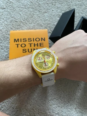 Omega x Swatch Mission To The Sun MoonSwatch - Säljer en Omega x Swatch MoonSwatch Mission to the Sun med gul boett och gul urtavla. Klockan har vita detaljer och vit textilrem med Omega-logga. Biokeramisk konstruktion och cool kronografdesign. Perfekt för dig som gillar färg och vill sticka ut.