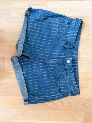 Blå randiga jeansshorts - Snygga blå jeansshorts med tunna vita ränder och uppvikta benslut. Klassisk femficksmodell med knapp och dragkedja framtill. Perfekta för varma dagar och enkel att matcha med olika toppar.