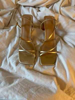 Guldiga sandaletter med smala remmar - Säljer ett par stilrena sandaletter från H&M i metallic guld med smala remmar och fyrkantig tå. Klackarna har en modern design och remmarna går snyggt runt foten. Perfekta för att lyfta vilken outfit som helst. Storlek 40 men passar även 39