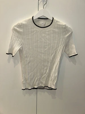 Vit ribbad topp med svarta kanter - Säljer en vit ribbad topp med svarta detaljer vid hals, ärmslut och nederkant. Toppen har korta ärmar och en kroppsnära passform som ger en fräsch och stilren look.