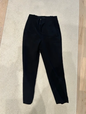 Svarta kostymbyxor med hög midja - Säljer ett par svarta kostymbyxor med hög midja och raka ben. Byxorna har fickor i sidorna och stängs med dragkedja och knapp. Perfekta till en snygg och clean look. Materialet känns mjukt och följsamt.