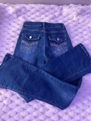 Mörkblå bootcut jeans med broderi - Säljer ett par mörkblå bootcut jeans med snygga broderade detaljer på bakfickorna och silverfärgade knappar. Jeansen har hög midja och klassisk femficksdesign. Perfekta för dig som gillar en retro vibe och vill sticka ut lite extra.