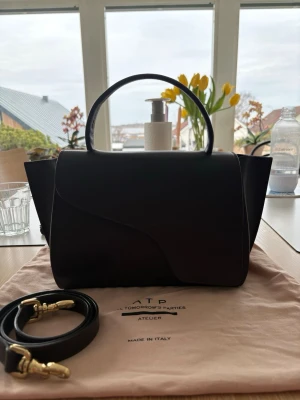 Mörkbrun handväska från ATP Atelier - Snygg mörkbrun handväska från ATP Atelier (Arezzo Vaccheta) i slätt läder. Väskan har ett stilrent, strukturerat formspråk med rundat handtag och avtagbar axelrem med guldfärgade detaljer. Tillverkad i Italien och perfekt för dig som gillar minimalistisk design. Är även köpt för 5200kr