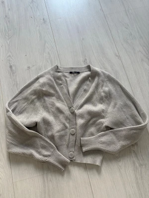 Grå  ribbad kofta med knappar - Grå kofta med ribbad struktur och stora knappar framtill. Kortare modell med v-ringning och långa ärmar. Perfekt att slänga över en topp för en chill look.