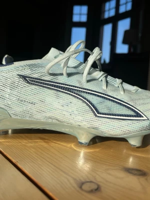 Ljusblå fotbollsskor från Puma  - Säljer ett par ljusblå fotbollsskor från Puma med vita och mörkblå detaljer. Skorna har snörning och en låg profil med mönstrad ovandel i syntetmaterial. Perfekta för dig som vill ha stilrena och lätta skor på planen. Skorna är använda ca 2-3 matcher och har dobbarna FG
