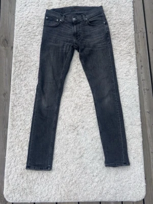 Gråa skinny jeans från Nudie Jeans - Säljer ett par gråa skinny jeans från Nudie Jeans med klassisk femficksmodell och svarta sömmar. Jeansen har en smal passform och är tillverkade i stretchigt denimtyg för extra komfort. Perfekta för en avslappnad och trendig look.