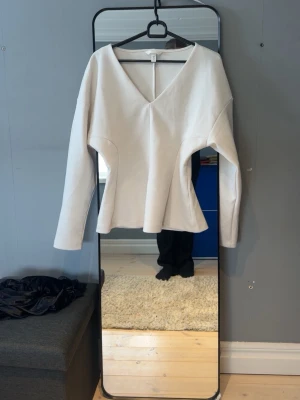 Vit v-ringad topp med lång ärm - Stilren vit topp med v-ringning och långa ärmar. Toppen har en lätt utsvängd passform och är tillverkad i ett mjukt material som känns bekvämt mot huden. Perfekt för att skapa en clean och modern look.