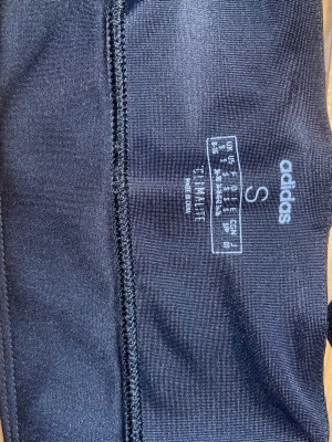Svart sport-bh från Adidas - Svart sport-bh från Adidas med vitt tryck framtill där det står 'adidas' och 'CODE OF SPORT'. Modellen har brottarrygg och är gjord i stretchigt funktionsmaterial (Climalite) som andas och transporterar bort fukt. Perfekt för träning och sport. Väl använd men också väl omhändertagen:)