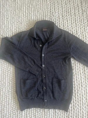 Mörkgrå cardigan från Oscar Jacobson - Stilren mörkgrå cardigan från Oscar Jacobson med knäppning framtill och två praktiska fickor. Tröjan har klassisk krage och är tillverkad i mjukt, tunt material som passar perfekt för lager på lager. Enkel och snygg design som funkar till det mesta.