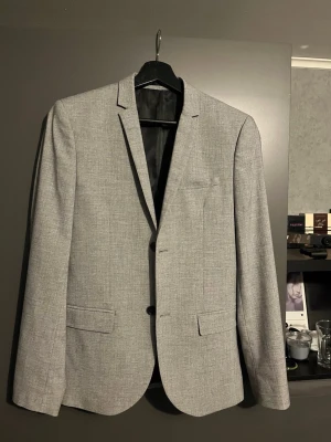 Ljusgrå kavaj med smal passform - Stilren ljusgrå kavaj med smal passform och klassisk design. Kavajen har två knappar framtill, två fickor med lock och en bröstficka. Tillverkad i ett tunt, vävt material med diskret struktur. Svart innerfoder och fyra knappar vid ärmslut.