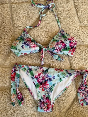 Blommig bikini med knyt i pastellfärger - Snygg bikini med blommigt mönster i grönt, rött, lila och vitt. Överdelen har knyt i nacken och framtill, medan underdelen har knyt i sidorna. Perfekt för sommarens strandhäng och poolparty.