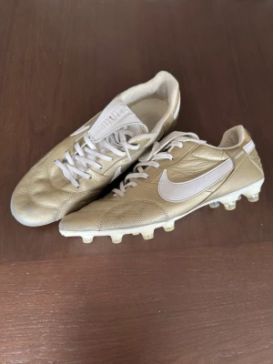  Nike Premier 3 FG - Snygga fotbollsskor från Nike Premier i guld och vitt. Skorna har klassisk snörning, vit sula med fasta dobbar och stort vitt Nike-märke på sidan. Ovandelen är i läder för bra känsla och passform. Perfekta för dig som vill sticka ut på planen.