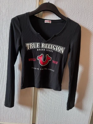 Svart  topp True Religion - Svart ribbad långärmad topp från True Religion med vit och röd logga och text framtill. Toppen har v-ringning och croppad passform, perfekt för en trendig streetwear-look.