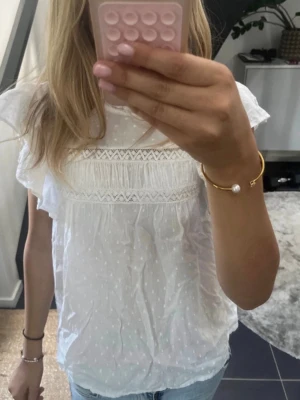Vit blus med spetsdetaljer - Söt vit blus med korta volangärmar och broderade spetsdetaljer över bröstet. Blusen har diskret prickmönster i tyget och en luftig, avslappnad passform. Perfekt till jeans eller kjol för en fräsch look.