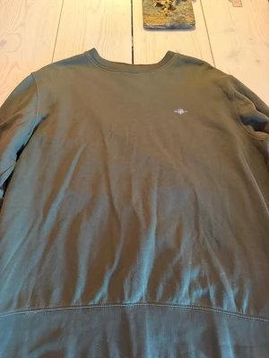 Olivgrön sweatshirt från GANT - Snygg olivgrön sweatshirt från GANT med rund halsringning och diskret logga broderad på bröstet. Tröjan har långa ärmar och är tillverkad i mjuk bomull, perfekt för en avslappnad stil.