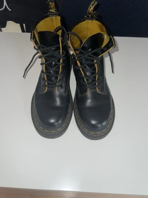 Svarta Dr. Martens kängor - Säljer ett par klassiska svarta Dr. Martens kängor med gul söm och högt skaft. Kängorna har snörning och är tillverkade i läder med gul insida och den ikoniska Dr. Martens-fliken