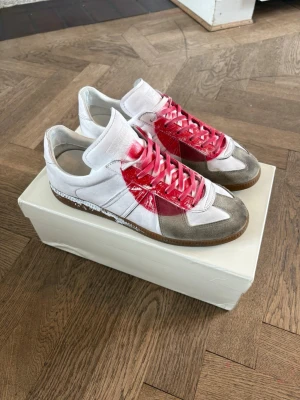 Maison Margeila Japan Gats - Unika sneakers Japan gats med vit bas, grå mockadetaljer och röd spray paint-effekt över snörning och sidor. Skorna har klassisk låg profil och bruna gummisulor. Materialet är en mix av läder och mocka, perfekt för dig som gillar statement pieces.