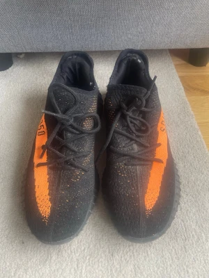 Yeezy Boost 350 V2 Black/Orange - Säljer ett par Yeezy Boost 350 V2 med svart Primeknit-överdel och en fet orange stripe med texten 'SPLY-350' på sidan. Skorna har en chunky sula och klassiska svarta skosnören. Perfekta för dig som vill sticka ut med en ikonisk sneaker.