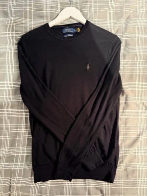 Svart tröja från Polo Ralph Lauren - Klassisk svart tröja från Polo Ralph Lauren med rund halsringning och diskret broderad logga på bröstet. Tillverkad i tvättbart merinoull, ett mjukt material som passar perfekt för lager-på-lager. Långa ärmar och ribbade muddar för en clean look.