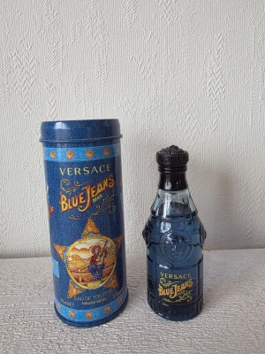 Versace Blue Jeans Man Eau de Toilette - Versace Blue Jeans Man Eau de Toilette med en unik blå glasflaska och tillhörande cylinderformad plåtburk med denim-inspirerat tryck och färgglada detaljer. Flaskan har klassisk Versace-design med reliefmönster och svart kork. Doften är fräsch och maskulin.
