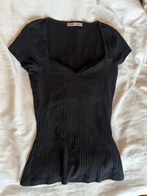 Svart ribbad topp från Anna Field - Snygg svart ribbad topp från Anna Field med v-ringad hals och korta ärmar. Toppen har en figurnära passform och är tillverkad i mjuk bomullsmix som känns skön mot huden. Perfekt att styla med jeans eller kjol för en trendig look. Storleken skulle jag säga är xs eller s 