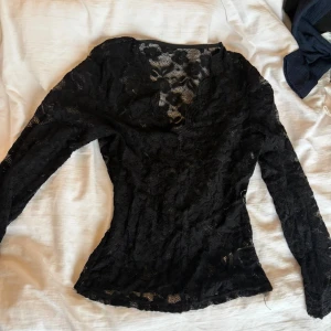 Svart spetsblus med v-ringning - Snygg svart blus i spets med v-ringad hals och långa ärmar. Transparent och figurnära modell som ger en elegant look. Perfekt att styla med jeans eller kjol för en trendig outfit.