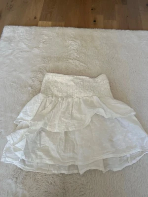 Vit volangkjol från H&M - Supersöt vit kortkjol från H&M  med flera lager volanger. Kjolen är gjord i ett lätt och luftigt bomullstyg, perfekt för varma dagar. Enkel att matcha med både toppar och sneakers för en chill look.