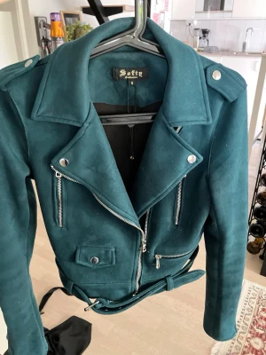 Mörkgrön biker skinnjacka med bälte - Snygg mörkgrön skinnjacka från Giorgio i klassisk bikerstil. Jackan har silvriga dragkedjor, axelklaffar, fickor med lock och ett matchande bälte i midjan. Perfekt för dig som vill ha en edgy look med coola detaljer.