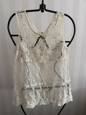 Vit spetslinne med dragkedja - Säljer ett vitt ärmlöst spetslinne med vackra virkade detaljer och transparenta partier. Toppen har rund halsringning och dragkedja i ryggen för en snygg look. Perfekt att styla med jeans eller kjol för en trendig outfit.