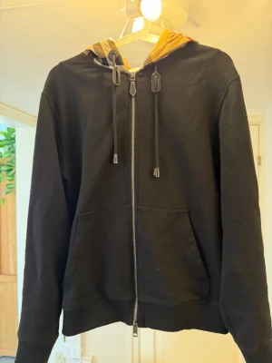 Burberry Hoodie - Svart hoodie med dragkedja och känguruficka. Huvan har ett snyggt rutmönster i beige, svart, vitt och rött, vilket ger en unik touch. Perfekt för dig som vill ha en stilren men ändå trendig look. Justerbara snören i huvan och mjukt material.