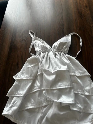 Vit volangig linnetopp i satin - Supersöt vit linnetopp med tunna axelband och flera lager volanger. Toppen har en v-ringad urringning och är gjord i glansigt satinliknande material som ger en lyxig känsla. Perfekt att styla med jeans eller kjol för en trendig look.❤️