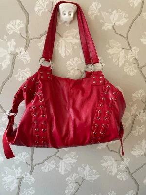 Röd handväska med metallringar - Cool röd handväska i konstläder med stora silverfärgade metallringar och dekorativa snörningar längs sidorna. Väskan har dubbla axelband och detaljer med nitar för en edgy look. Perfekt för dig som vill sticka ut med en färgstark accessoar.