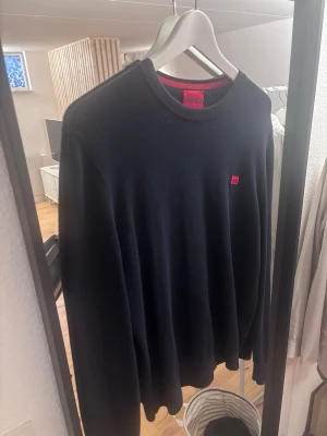 Svart stickad tröja från Hugo - Klassisk svart stickad tröja från Hugo med rund halsringning och diskret röd logga på bröstet. Tröjan har en enkel och stilren design, perfekt för dig som gillar minimalistiskt mode. Materialet känns mjukt och bekvämt. 