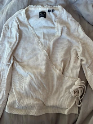 Vit omlottopp med spetsdetaljer - Supersöt vit omlottopp från superdry med ribbad struktur och fina spetsdetaljer längs ringningen. Långa ärmar och knytband i sidan ger en feminin look. Perfekt att styla med jeans eller kjol för en trendig outfit.