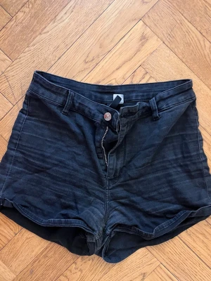 Svarta högmidjade jeansshorts - Snygga svarta högmidjade jeansshorts med klassisk femficksdesign och knappgylf. Shortsen har bälteshällor och bakfickor, perfekt för en avslappnad och trendig look. Passar till allt och är ett måste i garderoben.