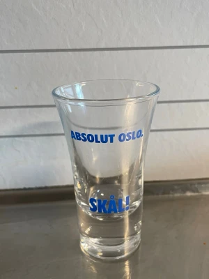 Shotglas med blå text Absolut Oslo - Säljer ett transparent shotglas med blå text som lyder 'ABSOLUT OSLO.' och 'SKÅL!'. Glaset har klassisk form och är tillverkat i glas, perfekt för att samla eller använda vid festliga tillfällen.