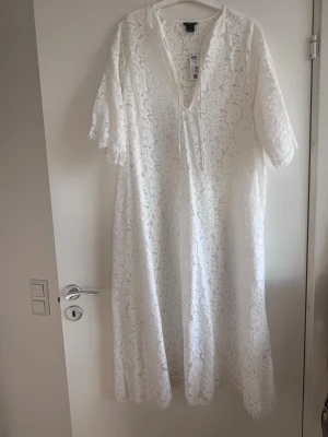 Vit spetsklänning från Lindex - Superfin vit långklänning från Lindex i spets med v-ringning och knytband fram. Klänningen har korta ärmar och ett blommigt spetsmönster som ger en romantisk vibe. Perfekt för sommarens alla tillfällen!
