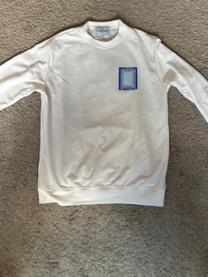 Vit sweatshirt från Casablanca - Vit sweatshirt från Casablanca med ett tryck i blått och grått på bröstet. Tröjan har rund halsringning, långa ärmar och ribbade muddar vid ärmslut och nederkant. Perfekt för en clean och stilren look. Den är i storlek S men är större i storleken. Skulle rekommendera för de som har M i storlek.
