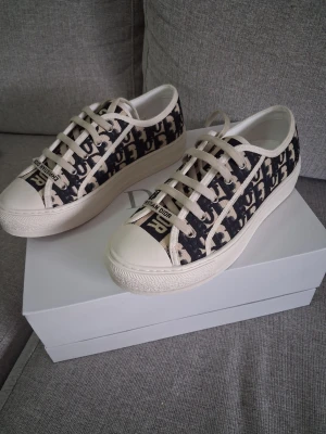 Dior Oblique låga sneakers - Snygga låga sneakers från Dior med klassiskt svart och vitt Oblique-mönster. Skorna har vita snören, vit sula och detaljer med 'CHRISTIAN DIOR' på tån. Tillverkade i canvas med gummisula, perfekta för en trendig streetstyle-look.