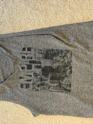 Grå ärmlös linnetopp med tryck - Snygg grå ärmlös linnetopp med mörkgrå tryck av stadsmotiv och texten 'EMPIRE' på framsidan. Toppen har en avslappnad passform och är tillverkad i mjukt material, perfekt för en cool och urban stil. Passar med både jeans och mjukisbyxor.