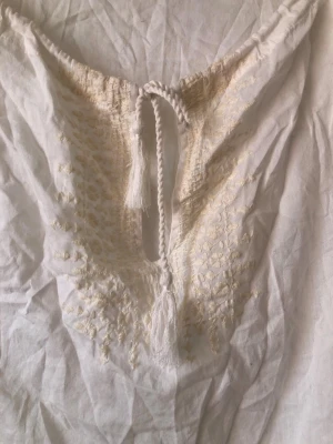 Vit bohemisk blus med broderi - Vit romantisk blus med bohemisk känsla, broderade detaljer vid halsringningen och ärmsluten samt knytning med tofs framtill. Tillverkad i ett lätt och luftigt tyg, perfekt för en avslappnad och stilren look. Resårbandet i midjan är något snott, går ev rätta till. Syns ej mycket.