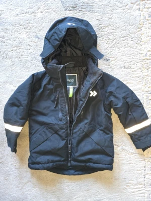 Svart vindjacka från Five Seasons - Svart vindjacka med huva från Five Seasons. Jackan har dragkedja, två fickor framtill och reflekterande band på ärmarna. Perfekt för blåsiga dagar och har en sportig look med logga på bröstet.