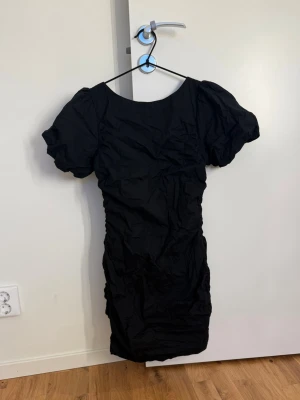 Svart kort puffärmad klänning - Snygg svart kortklänning med puffärmar och rynkade detaljer. Klänningen har en rund halsringning och är tillverkad i ett lätt, syntetiskt material. Perfekt för dig som vill ha en enkel men trendig look.