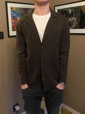 Mörkbrun stickad kavajtröja från John Henric  - Snygg mörkbrun stickad kavajtröja med tre knappar framtill och tre fickor. Tröjan har en avslappnad passform och klassisk krage, perfekt att slänga över en t-shirt för en clean look. Materialet ser mjukt och bekvämt ut.