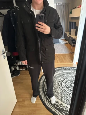 Parkajacka - Säljer en svart parkajacka med hög krage och flera praktiska fickor framtill. Jackan har dragkedja och knappar, samt en stilren och enkel design som passar till det mesta. Perfekt för kyligare dagar och har en normal passform.