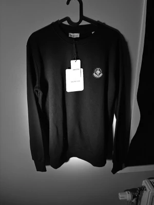 Svart sweatshirt från Moncler - Snygg svart sweatshirt från Moncler med rund halsringning och Moncler-logga på bröstet. Tröjan har långa ärmar och en klassisk, stilren design som passar perfekt till jeans eller joggers. Materialet känns mjukt och bekvämt.