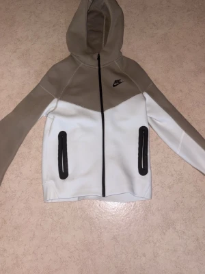 Nike beige och vit hoodie - Nike hoodie i beige och vitt med dragkedja framtill och två svarta fickor. Hoodien har en snygg tvåfärgad design och Nike-logga på bröstet. Tillverkad i mjukt material som passar perfekt till en avslappnad streetwear-look.