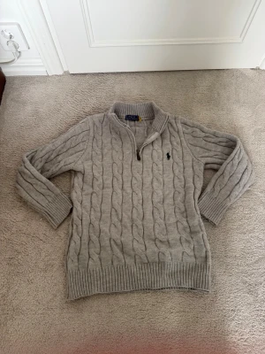 Grå kabelstickad tröja Polo Ralph Lauren - Kabelstickad grå tröja från Polo Ralph Lauren med halv dragkedja och broderad logga på bröstet. Tröjan har ribbade muddar och krage, perfekt för lager-på-lager. Klassisk och stilren design som funkar till det mesta.