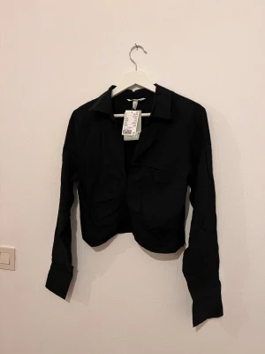 Skjorta/magtröja, helt ny! Slutsåld. - Magtröja från H&M i storlek M. Superfint material och så snygg på! Passar tyvärr inte mig. Har knappar där framme för att stänga så man kan ha den öppen ifall man vill. Men när man knappar upp den så syns inte knapparna 😊 Den har en krage och är lite krinklat vid bröstet! Svårt att ta bild på svarta plagg men det är bara att höra av sig ifall du har en fråga!